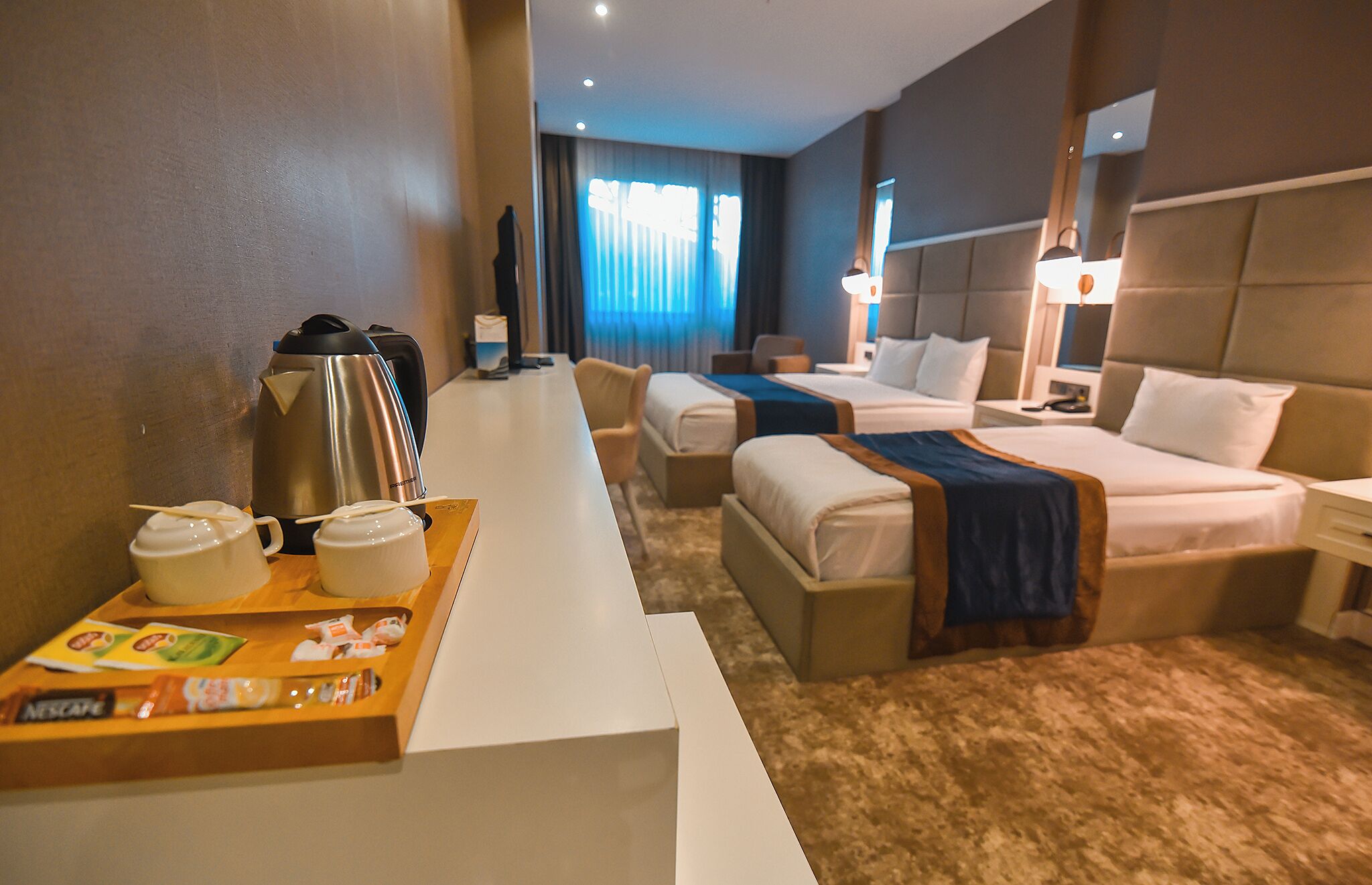 A hotel room with two beds, a minibar, a coffee maker, and a desk with a tray of breakfast items.