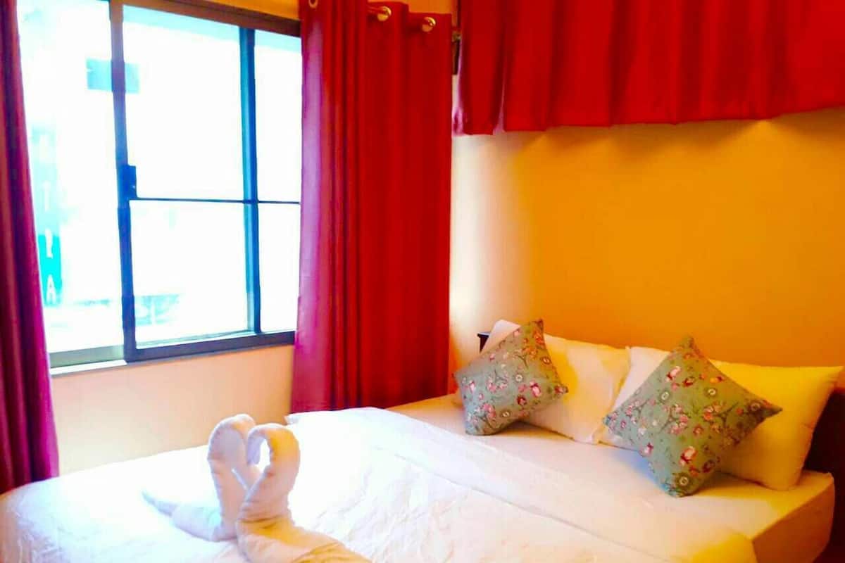 Un lit bien fait, avec du linge de lit blanc et des coussins décoratifs, une fenêtre ornée de rideaux rouges et un mur jaune aux tons chaleureux.