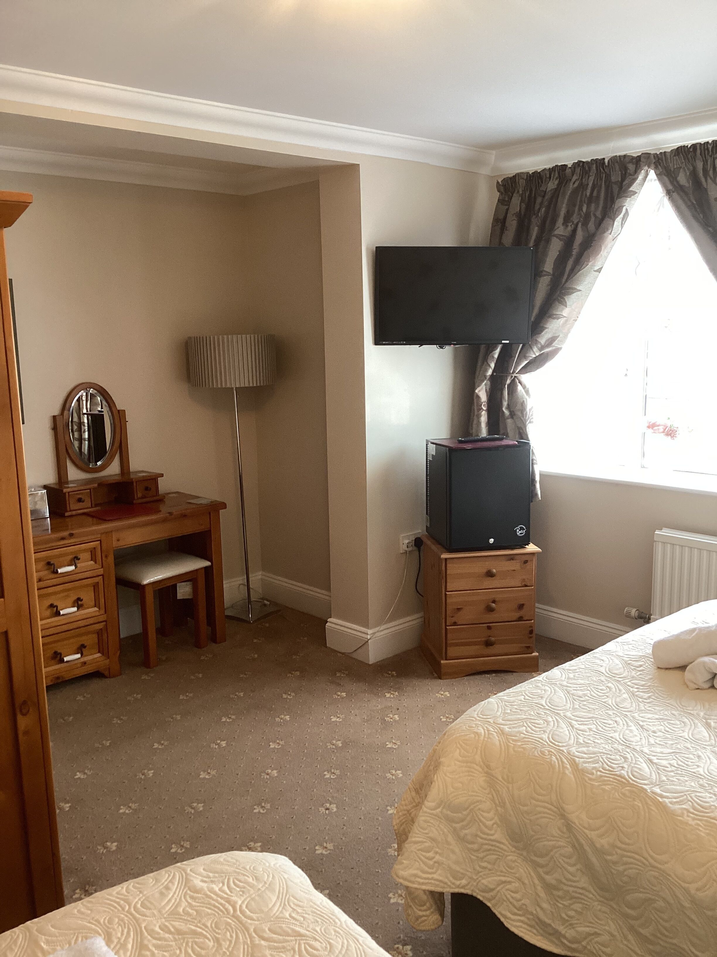A hotel room with a bed, a desk with a mirror, a television on a stand, a lamp, and a window with curtains.