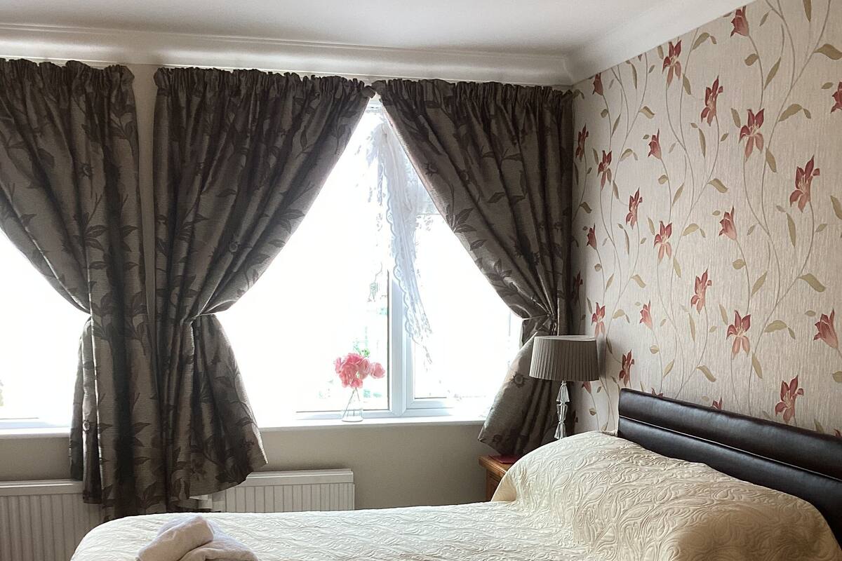 A hotel room with two beds, a window with curtains, a lamp, and a floral wallpaper.
