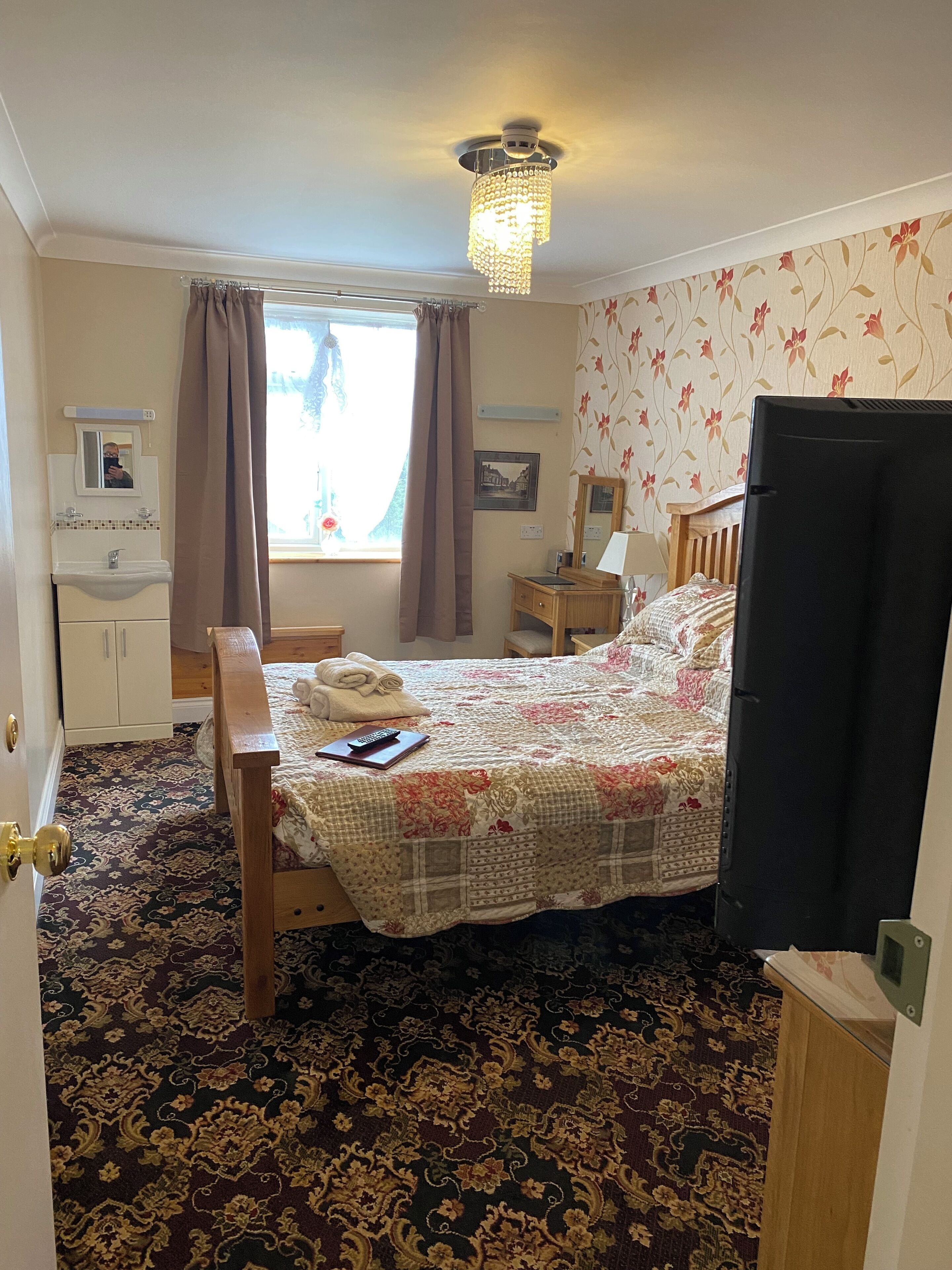 A bedroom with a bed, a wooden dresser, a desk, a mirror, a window with curtains, and a chandelier.