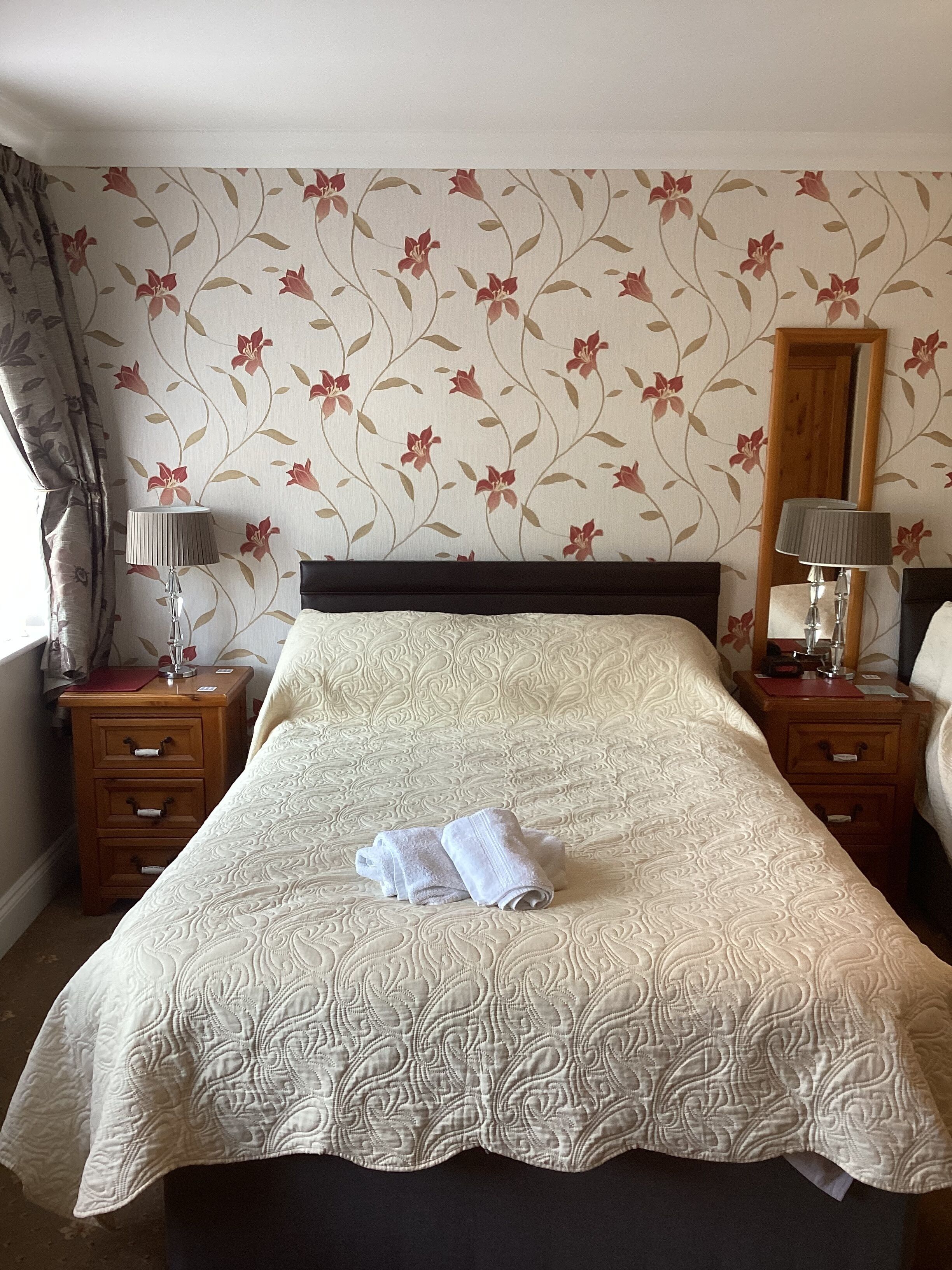 Una cama bien hecha con un papel tapiz estampado con motivos florales, dos mesitas de noche con lámparas, y un espejo en la pared.