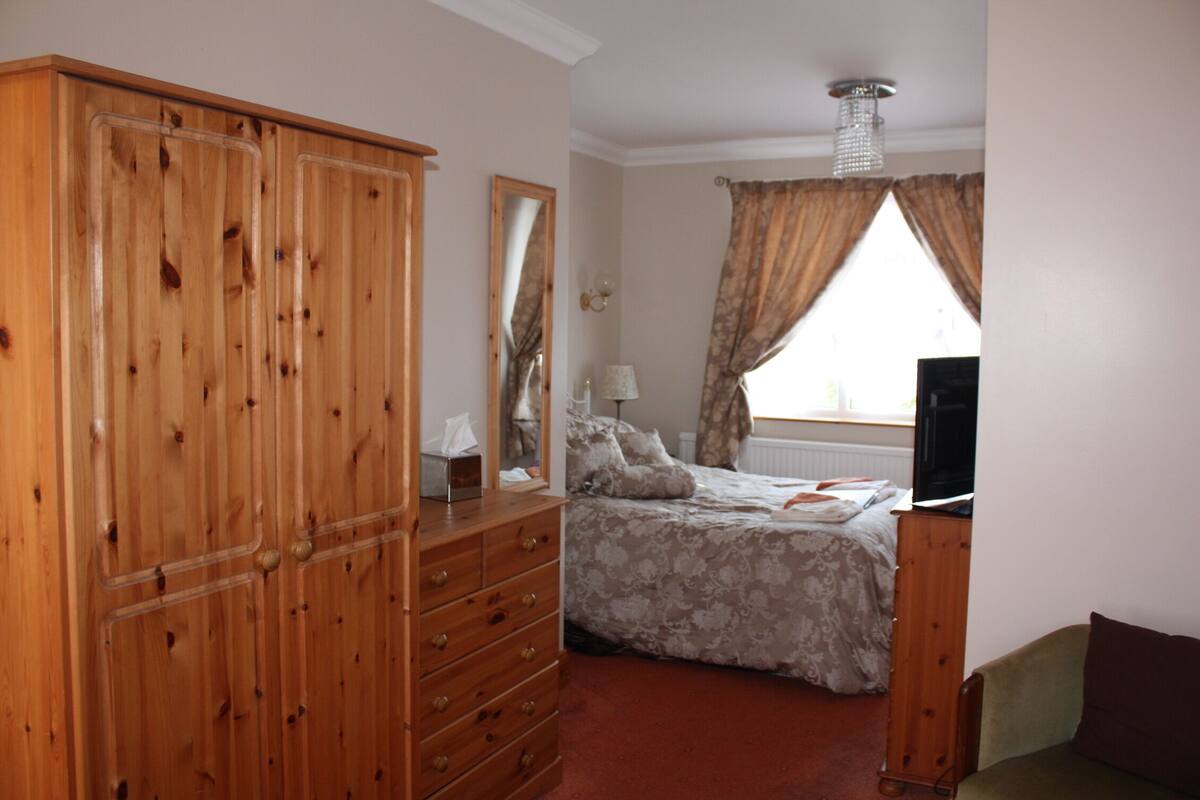 A bedroom with a wooden wardrobe, a bed with a floral duvet, a bedside table with a lamp, and a television on a stand.