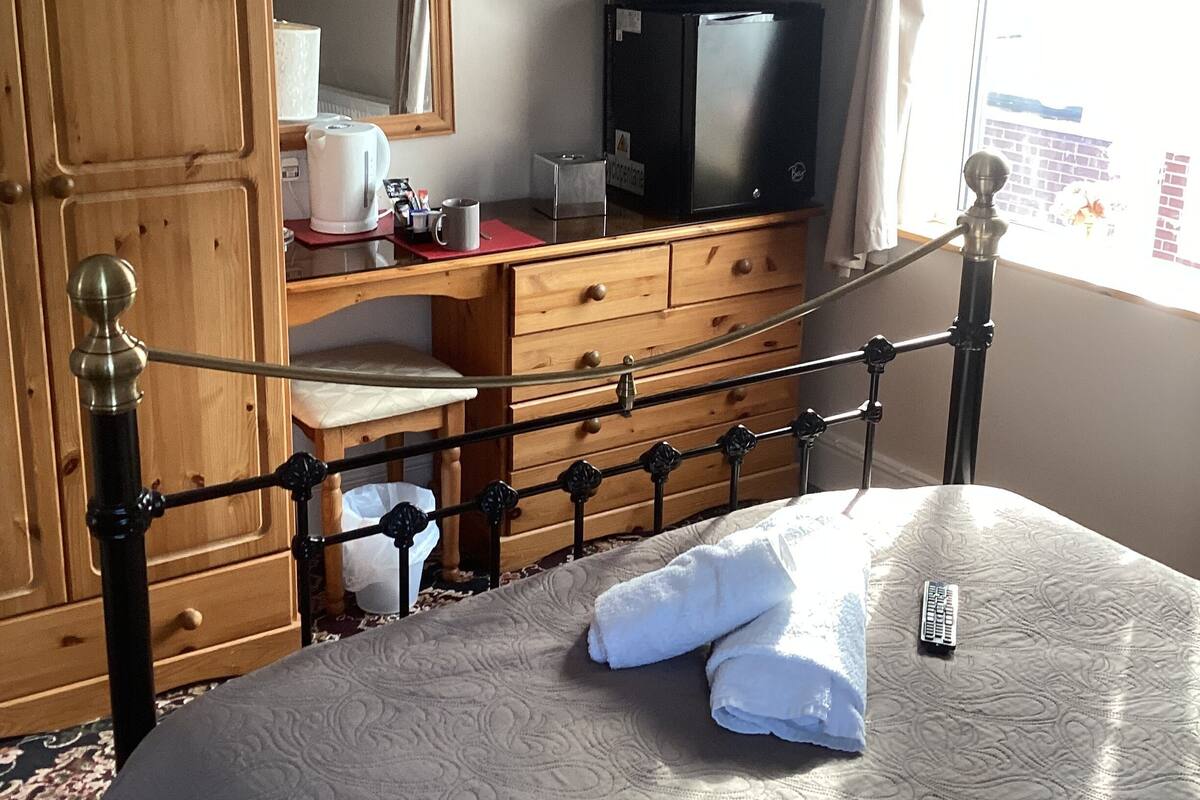 A bedroom with a bed, a wooden wardrobe, a desk with a television, and a window with curtains.