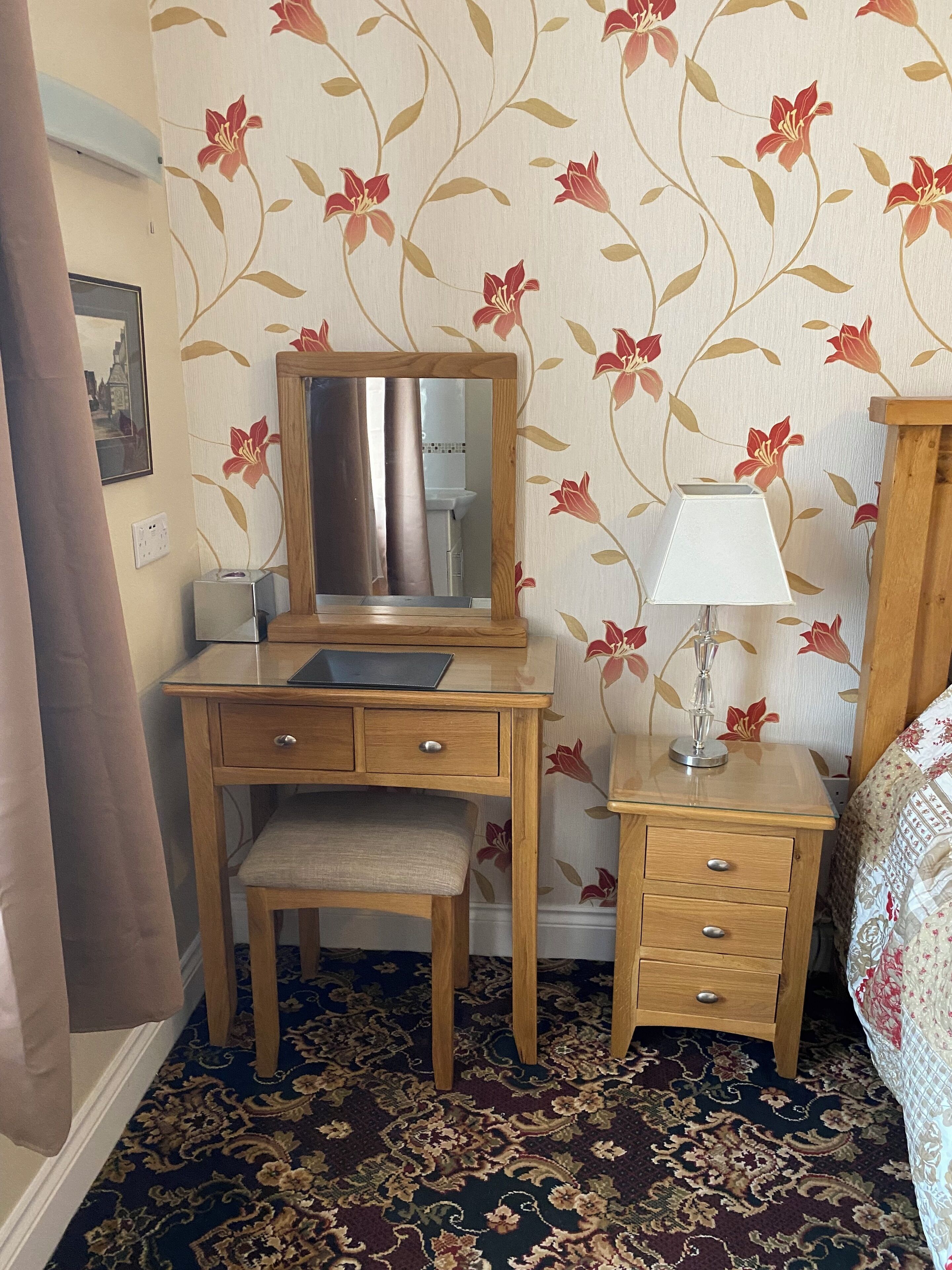 A small, neatly arranged bedroom with a wooden desk, a mirror, a bedside table with a lamp, and a bed with a patterned bedspread.