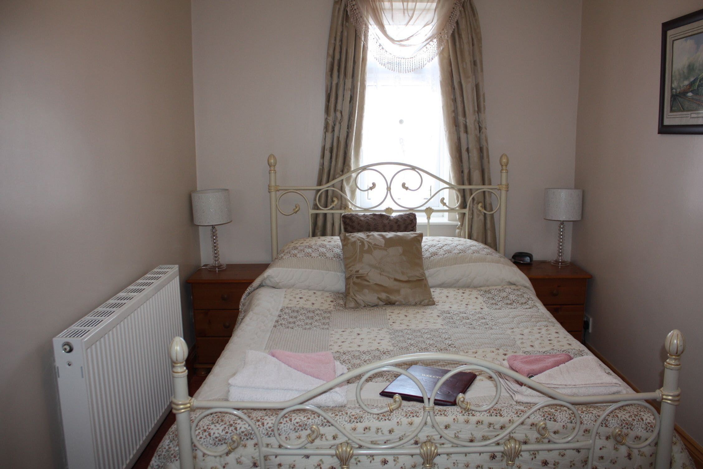 A neatly made bed with a floral-patterned bedspread and pillows, flanked by two bedside tables with lamps, a radiator, and a window with curtains.