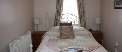 A neatly made bed with a floral-patterned bedspread and pillows, flanked by two bedside tables with lamps, a radiator, and a window with curtains.