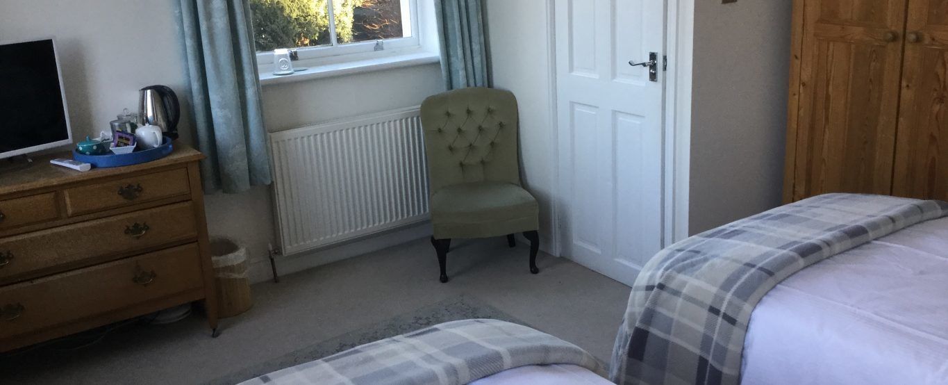 Une chambre à coucher comprenant un lit, une commode en bois, une chaise, un radiateur, une fenêtre avec des rideaux et une porte.