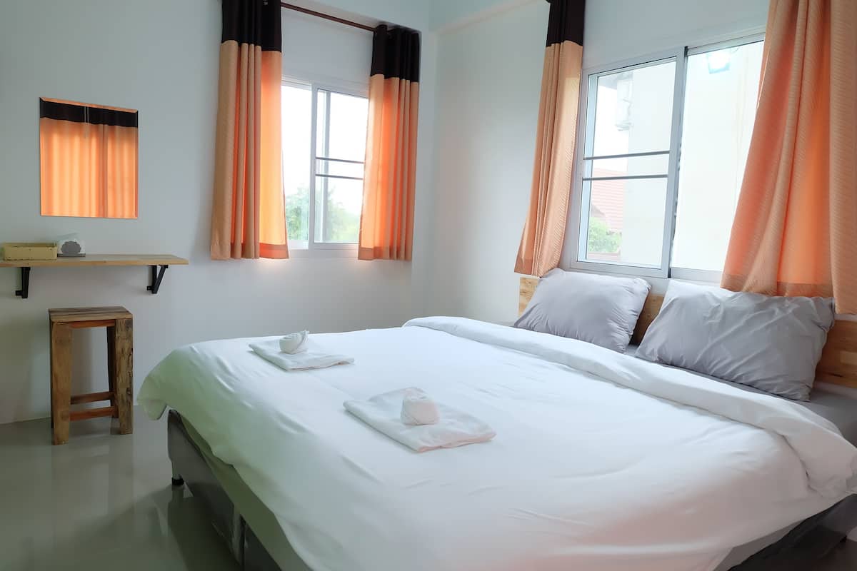 A neatly made bed with white linens and two folded towels, a wooden nightstand, a small table with a lamp, and two windows with orange curtains.