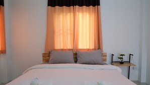 Ein ordentlich bezogenes Bett mit weißen Leinen und zwei Handtüchern, ein kleiner Nachttisch mit einem Topf und einer Pflanze sowie ein Fenster mit orangefarbenen Vorhängen.