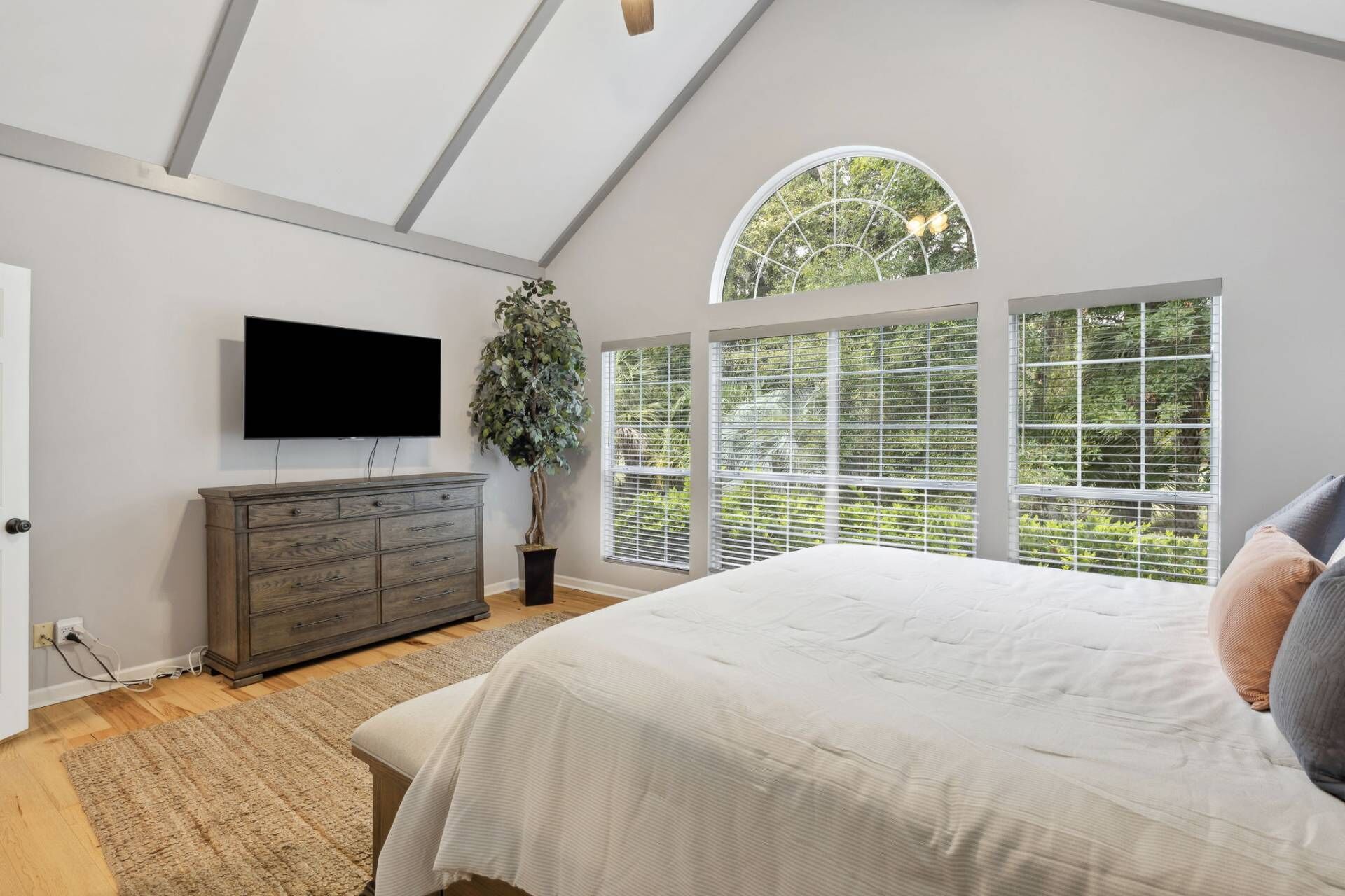 Un dormitorio con cama, cómoda, televisor y ventanas amplias con persianas.