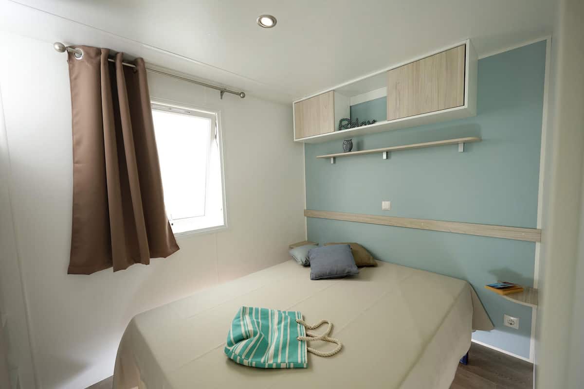 Une chambre à coucher avec un lit, une fenêtre avec des rideaux, une étagère ornée d’un objet décoratif et une petite table avec des livres.