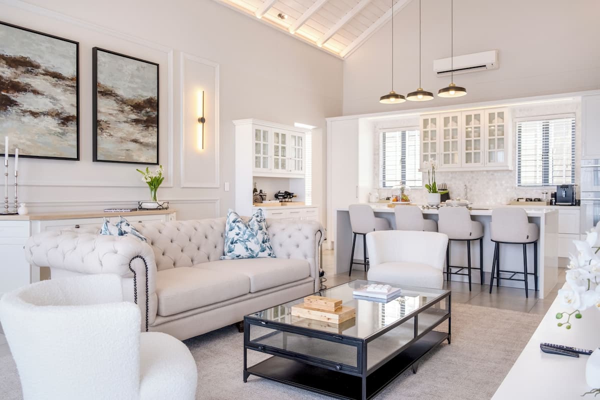 Una sala de estar moderna con un sofá, sillones y una mesa de centro de vidrio. Incluye una zona de cocina con taburetes de bar y una mesa de comedor.