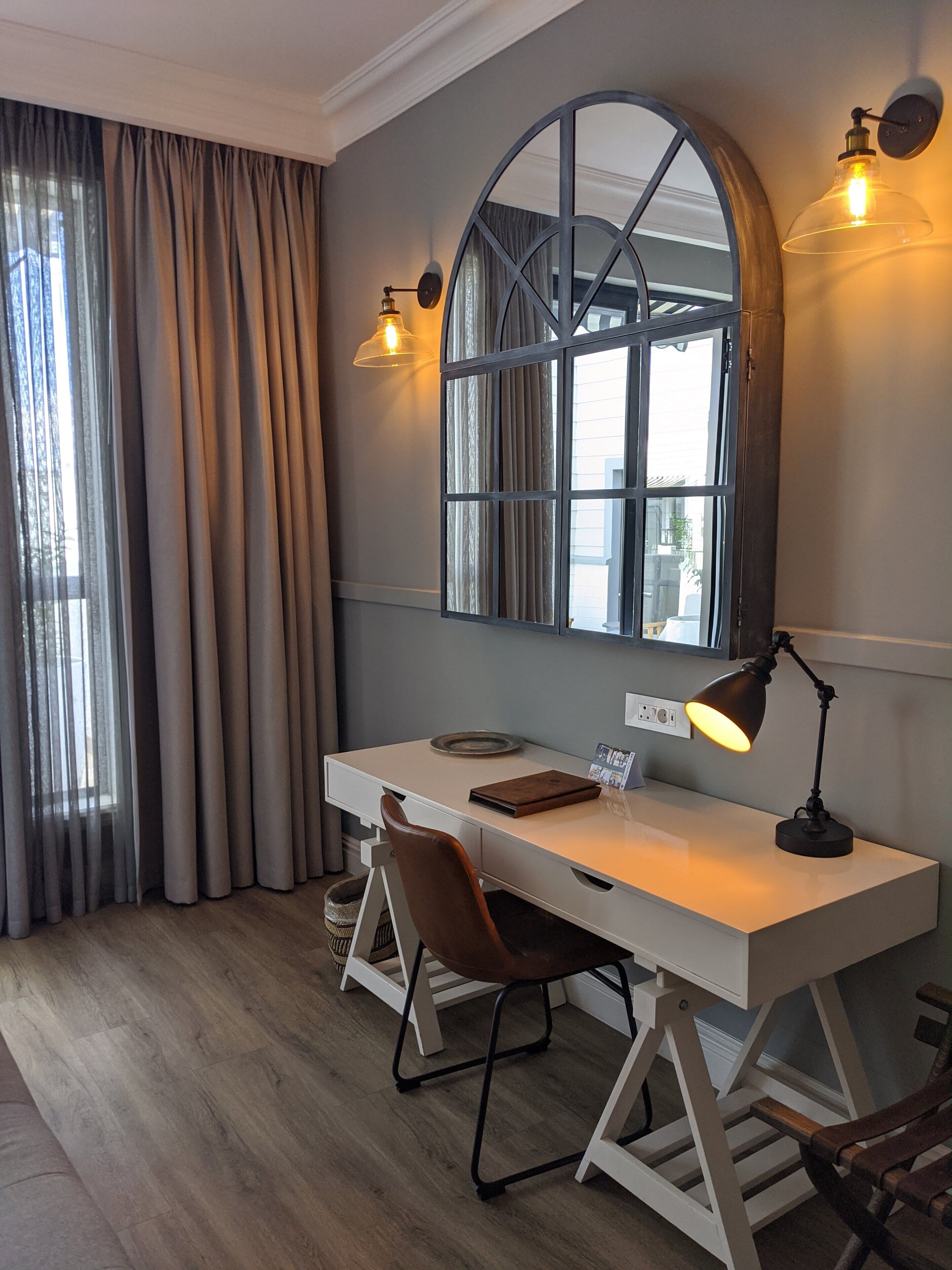 A modern office space with a white desk, two chairs, a large mirror, and a desk lamp.
