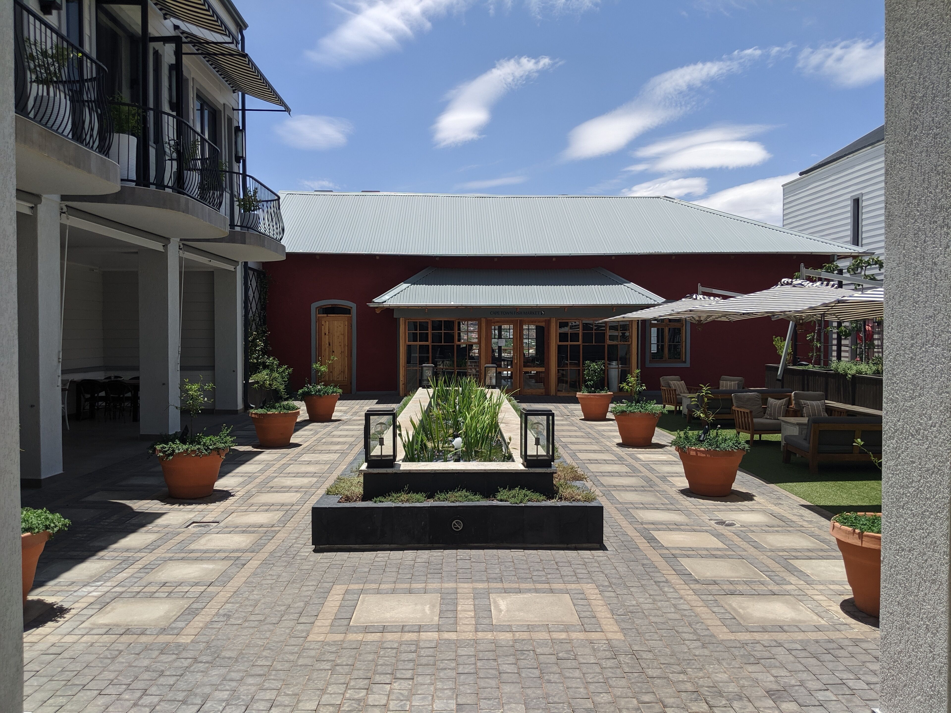 A courtyard with a central planter, potted plants, outdoor seating, and a building with a red facade and a metal roof.