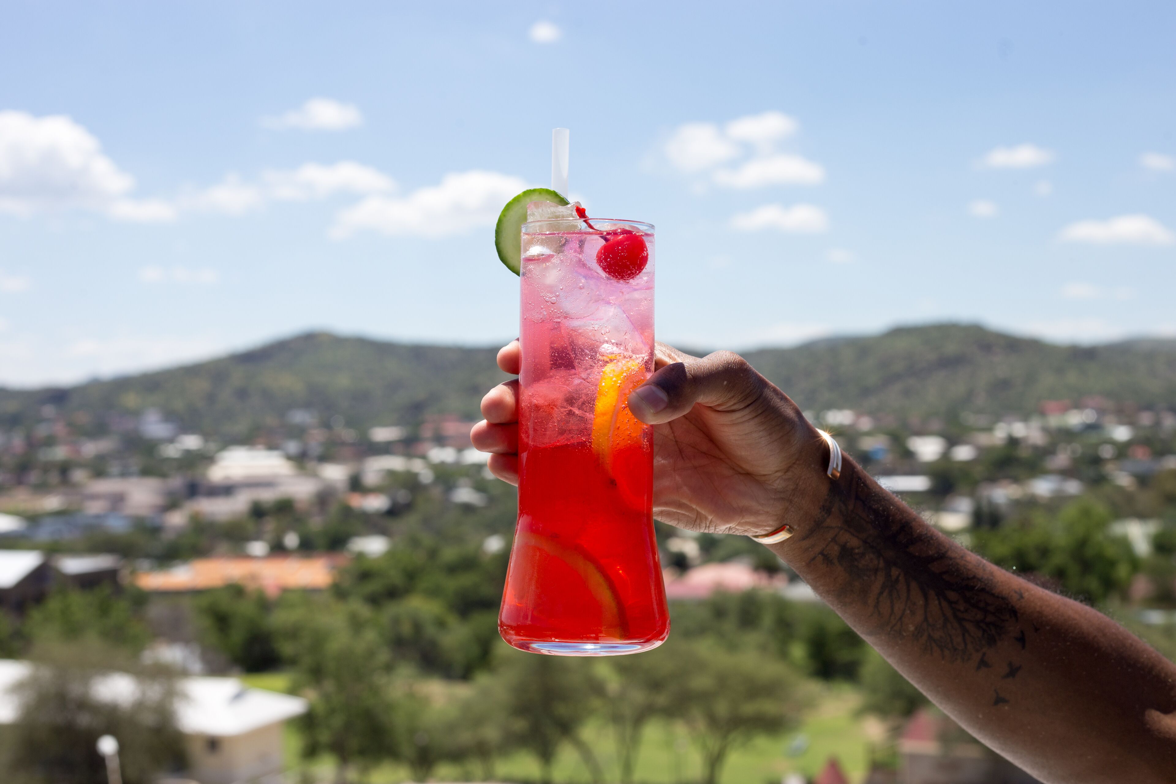 Een hand houdt een hoog glas met een rood drankje vast, met een partje citroen en een kers, voor een achtergrond van een stad en bergen.
