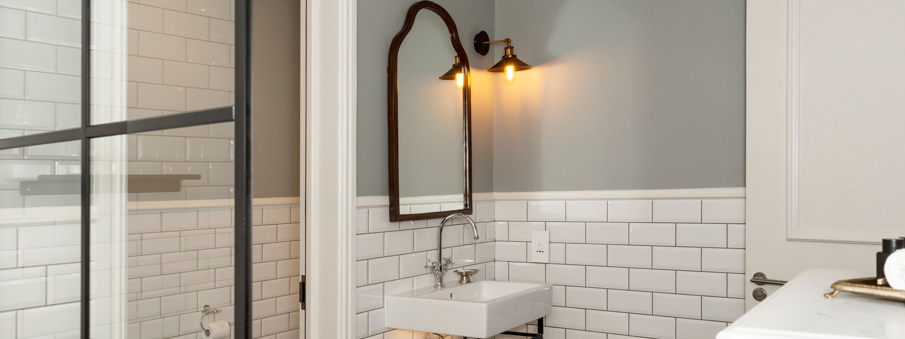 Une salle de bain avec des murs recouverts de carreaux blancs, un lavabo blanc, un miroir avec un cadre en bois et deux appliques murales.