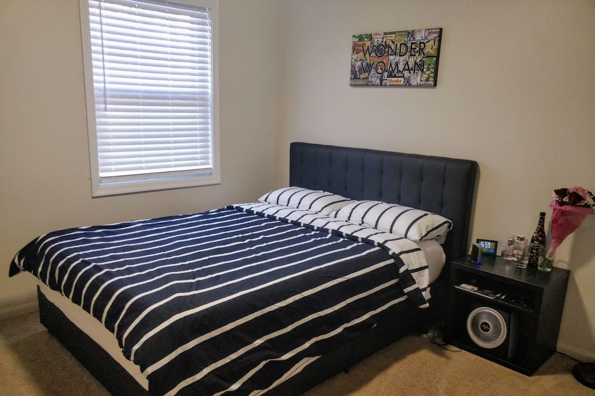 A bedroom with a bed, a nightstand, a wall clock, and a window with blinds.