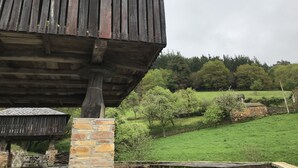 Eine rustikale Holzhütte mit Steinfundament, vermutlich ein Kiosk oder Pavillon, eingebettet in eine üppige grüne Landschaft mit Bäumen und einer Steinmauer im Hintergrund.