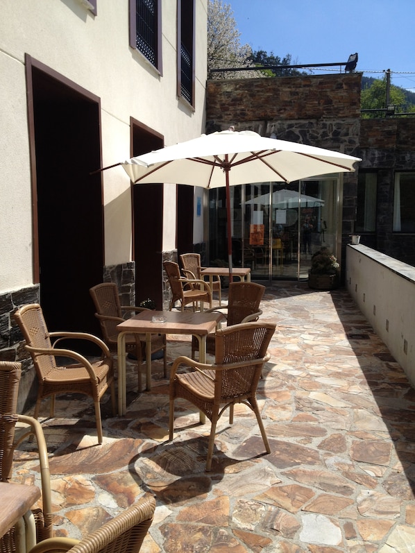 Eine Terrasse im Freien mit Korbmöbeln, einem großen Sonnenschirm, Steinböden und Blick auf ein Gebäude mit einer steinernen Fassade.