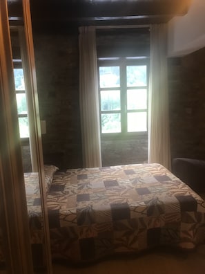 Ein Zimmer mit einem Bett, einem Fenster mit Vorhängen und Blick auf Grün außerhalb.