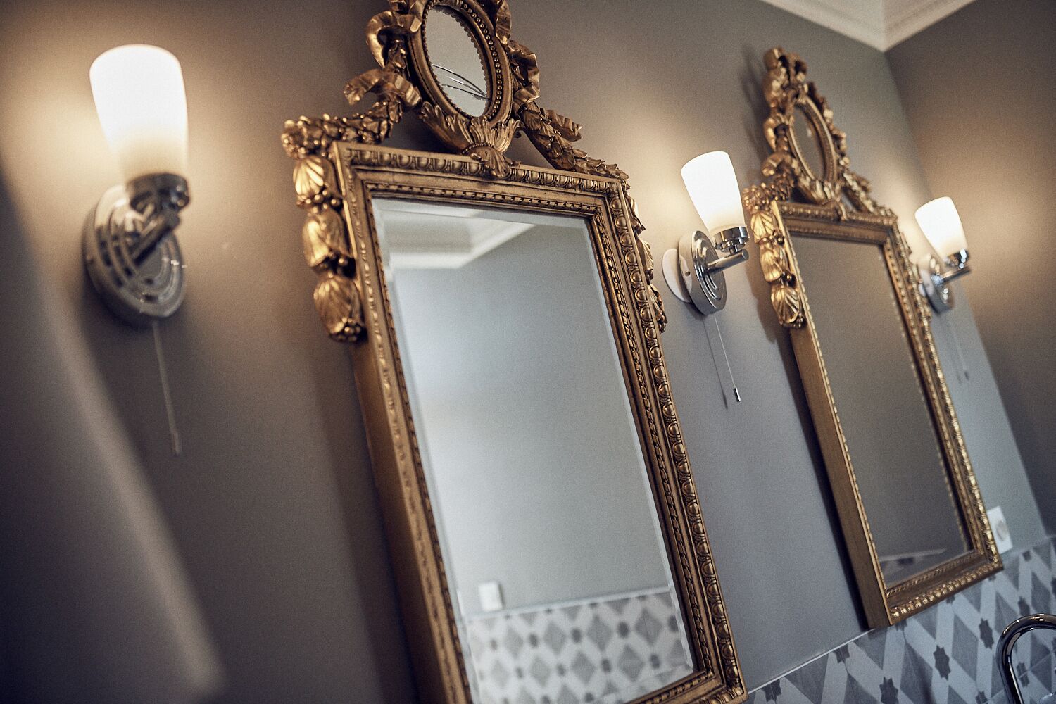 Two ornate mirrors with wall sconces, a patterned tile backsplash, and a towel rack.