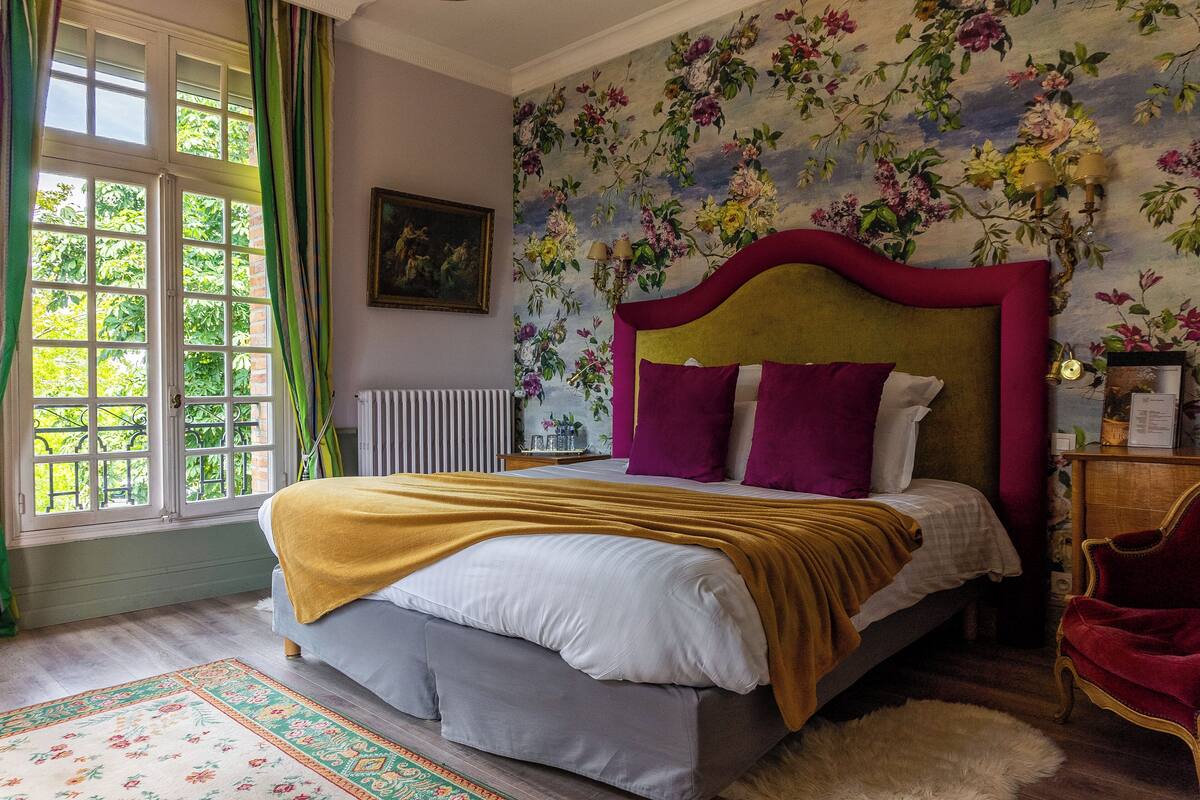 Ein Schlafzimmer mit einem großen Bett, floralem Wandpapier, einem Fenster mit Vorhängen und einer Heizung.