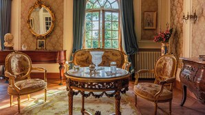 A room with a large ornate mirror, a round table with a glass top, two upholstered chairs, a wooden dresser, and a window with curtains. - Castel Bois Marie (Montauban)