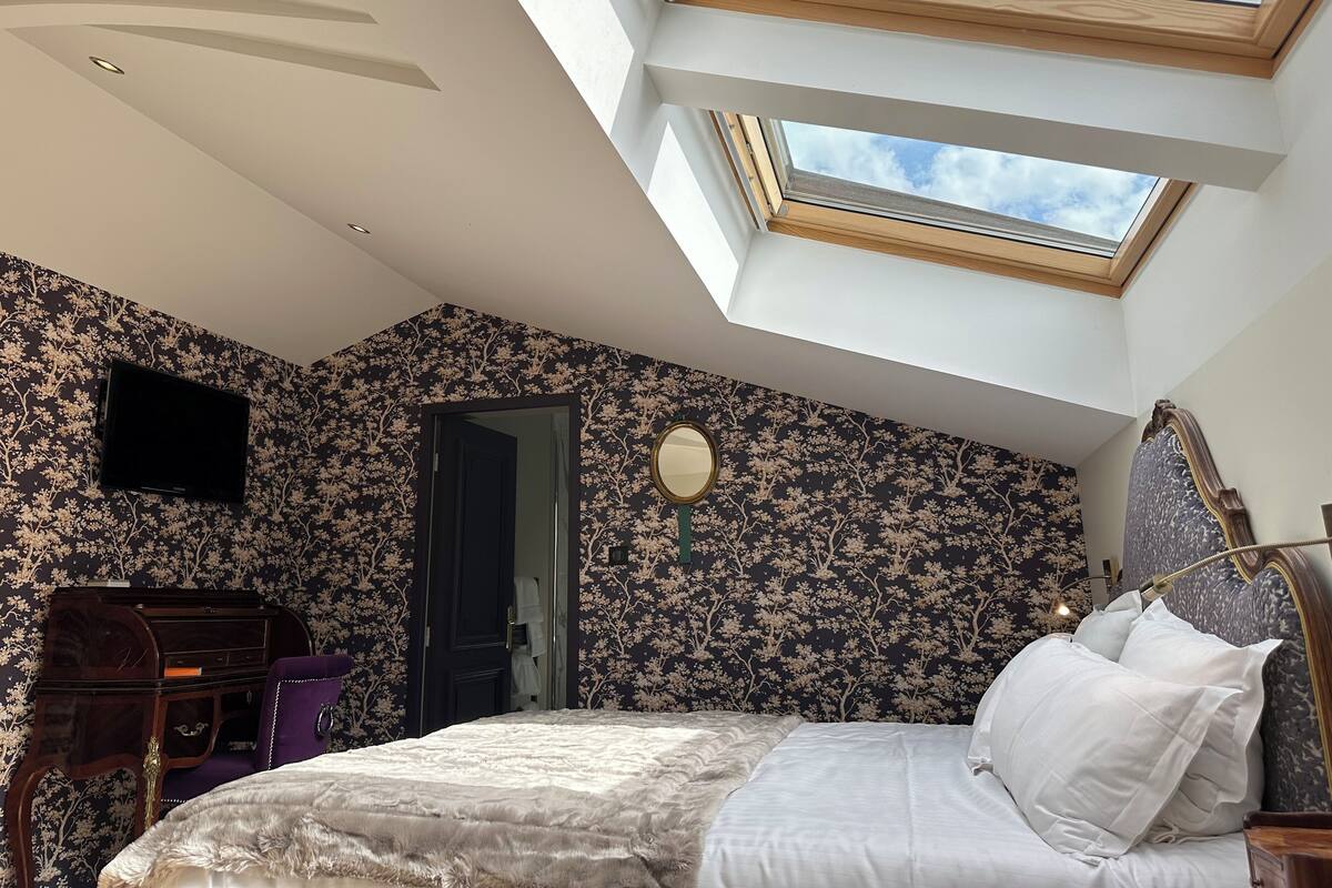 Ein Schlafzimmer mit Oberlicht, einem Bett mit weißer Bettwäsche, einem Schminktisch mit Spiegel und floralem, dunklem Tapetenmuster.