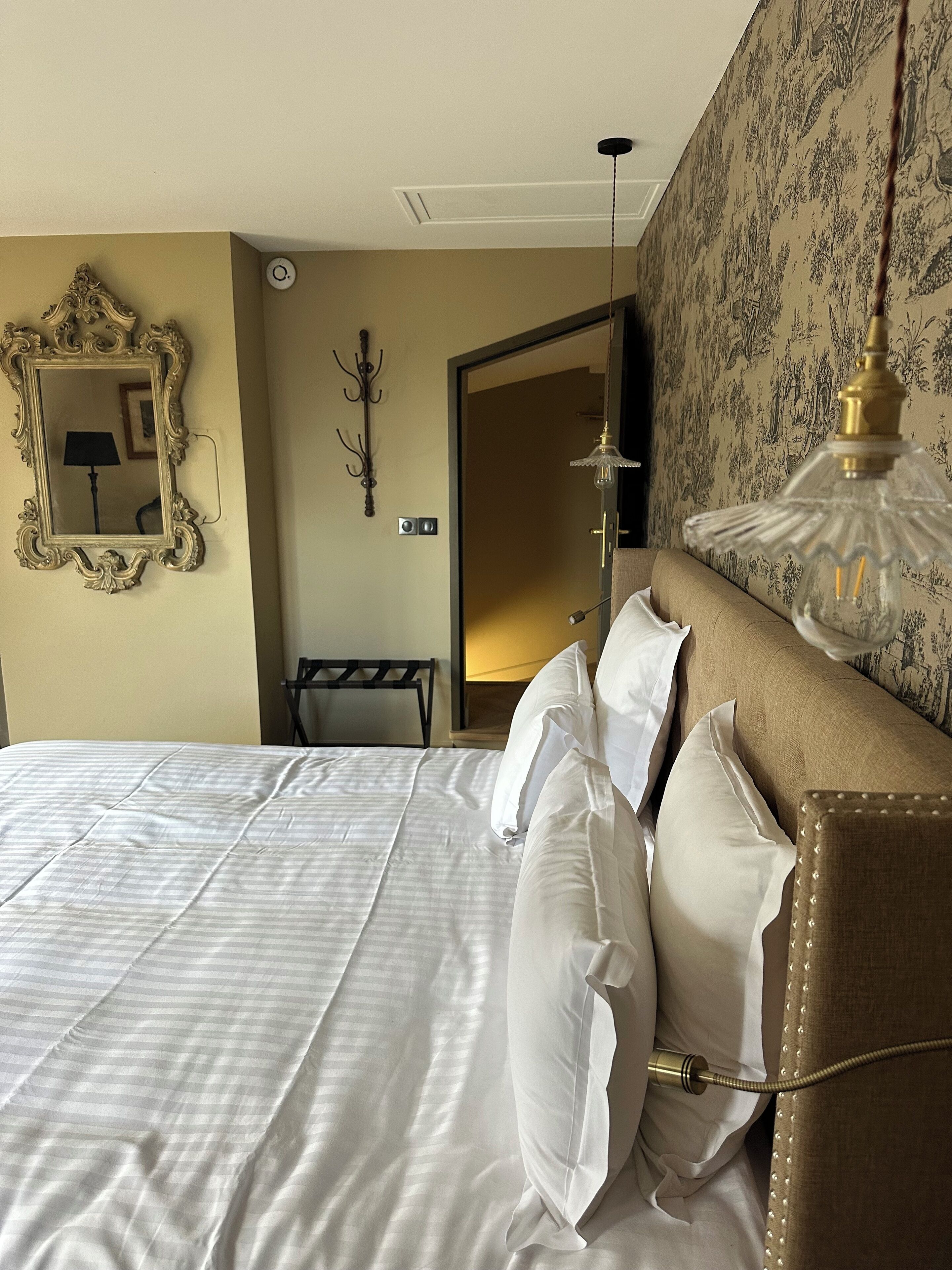A neatly made bed with white linens, a large mirror with an ornate frame, a wall-mounted lamp, and a door leading to another room.