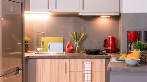 A modern kitchen with a stainless steel refrigerator, wooden cabinets, a sink, a cutting board, a red kettle, a frying pan, and a bowl of fruit. - Eternal luxury suites (Athens)