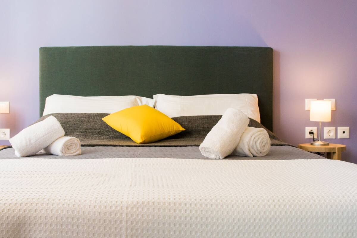 A neatly made bed with a green headboard, white and gray bedding, a yellow pillow, and two rolled towels. There are two bedside lamps on wooden nightstands.