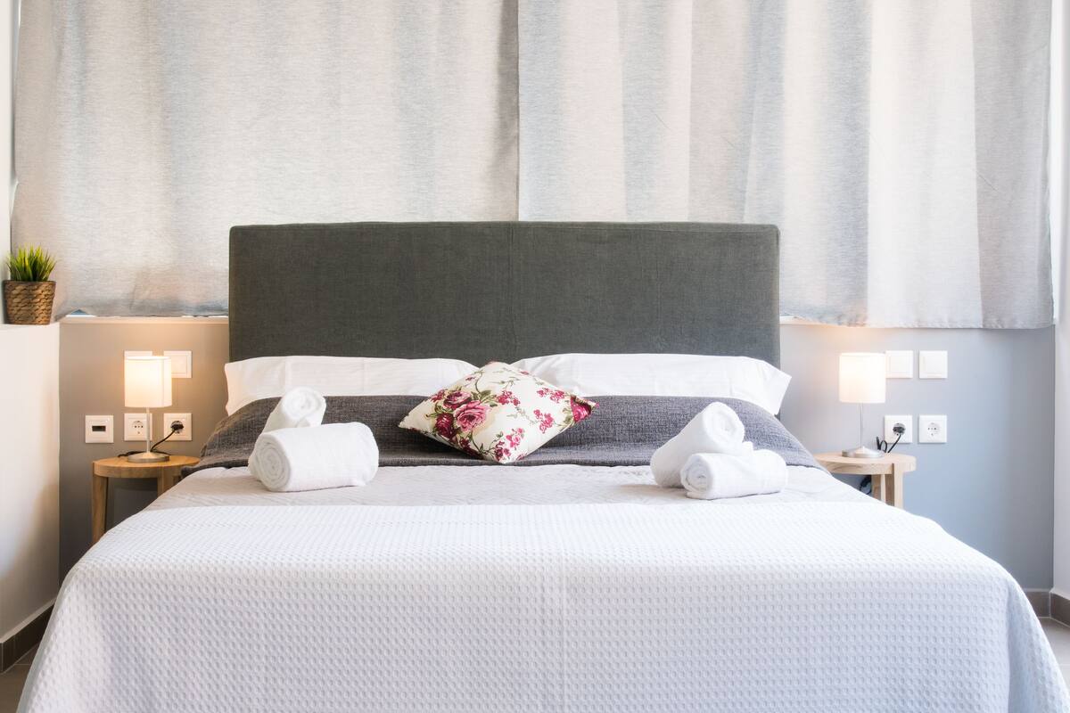A neatly made bed with white bedding, a floral pillow, and rolled towels. There are bedside lamps, a small plant, and a wall-mounted power outlet.