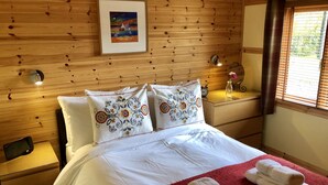 Una habitación con paneles de madera, una cama, almohadas, una mesita de noche, una lámpara y un ventanal con persianas.