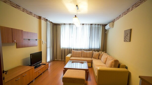 A living room with a sofa, ottoman, TV, and wooden cabinet. - Apartment Geo Milev (Plovdiv)