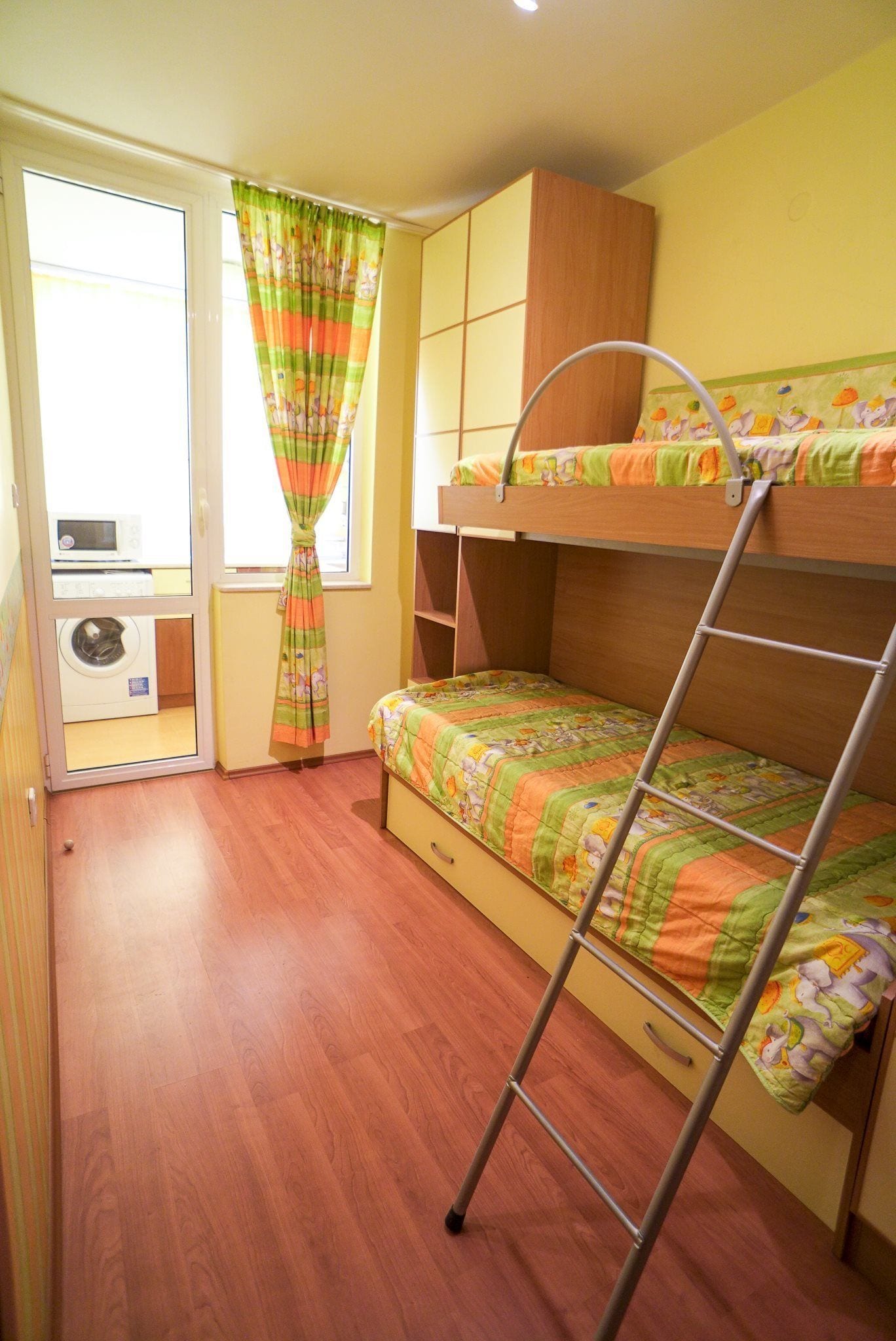 Ein Schlafsaal mit Etagenbetten, einem Holzschrank, einem kleinen Fenster mit Vorhängen und einer Waschmaschine in der Ecke.