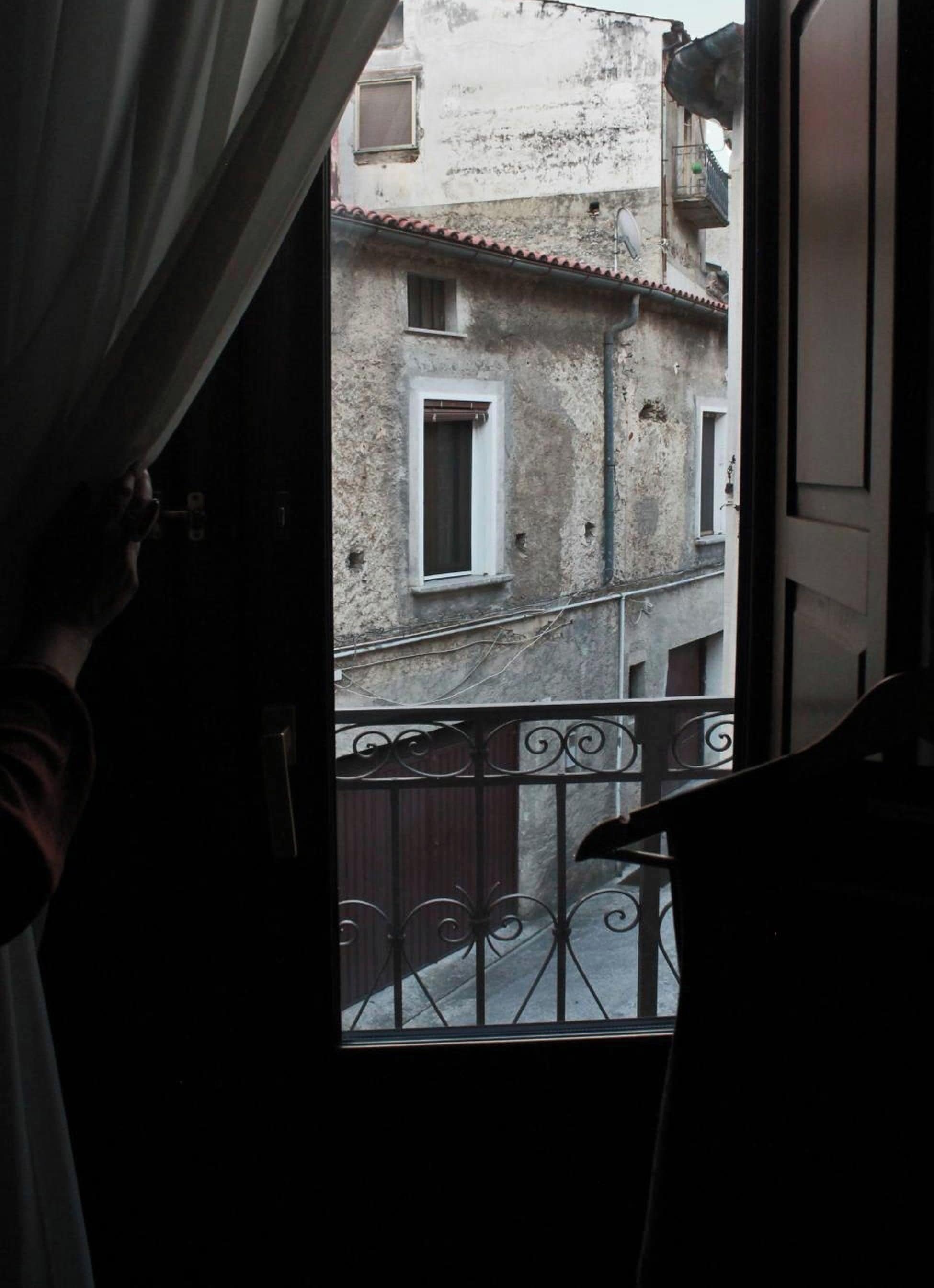 A view from a balcony with a metal railing, a building with multiple windows, and a curtain partially drawn to the side.