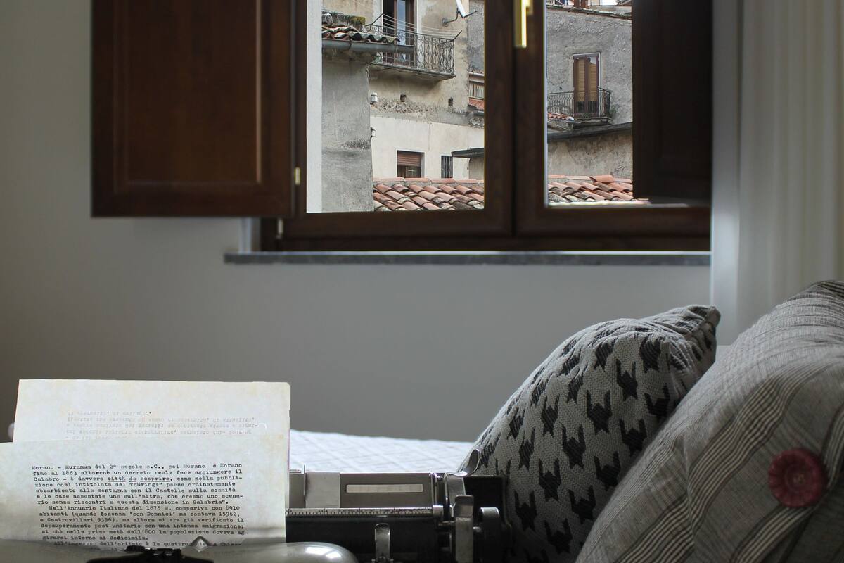 A room with a view of buildings through a window, a typewriter, a book, and a pillow with a pattern.