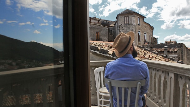 A man sitting on a balcony chair overlooking a historic town with stone buildings and a mountainous backdrop.