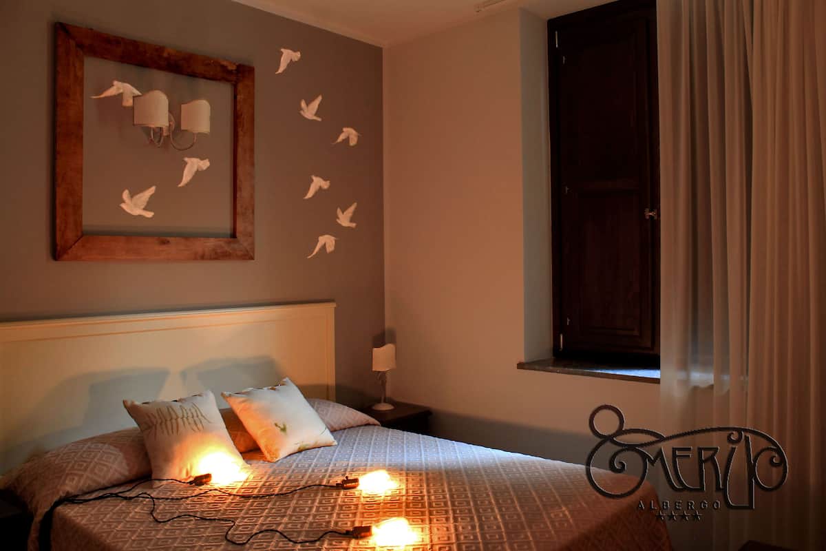 A bedroom with a patterned bedspread, two pillows, a bedside lamp, and a framed artwork with paper birds on the wall.