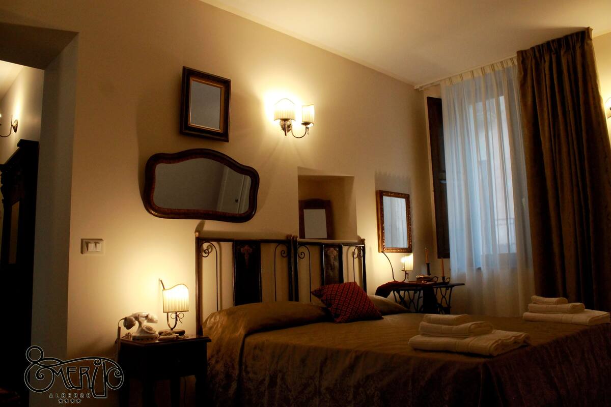 A hotel room with a bed, a bedside table with a lamp, a mirror, and a window with curtains.