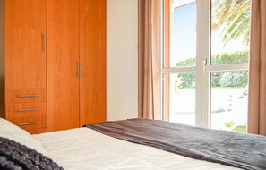 Un dormitorio con un armario de madera, una cama con colchón blanco y manta marrón, y una ventana con vista a follaje.