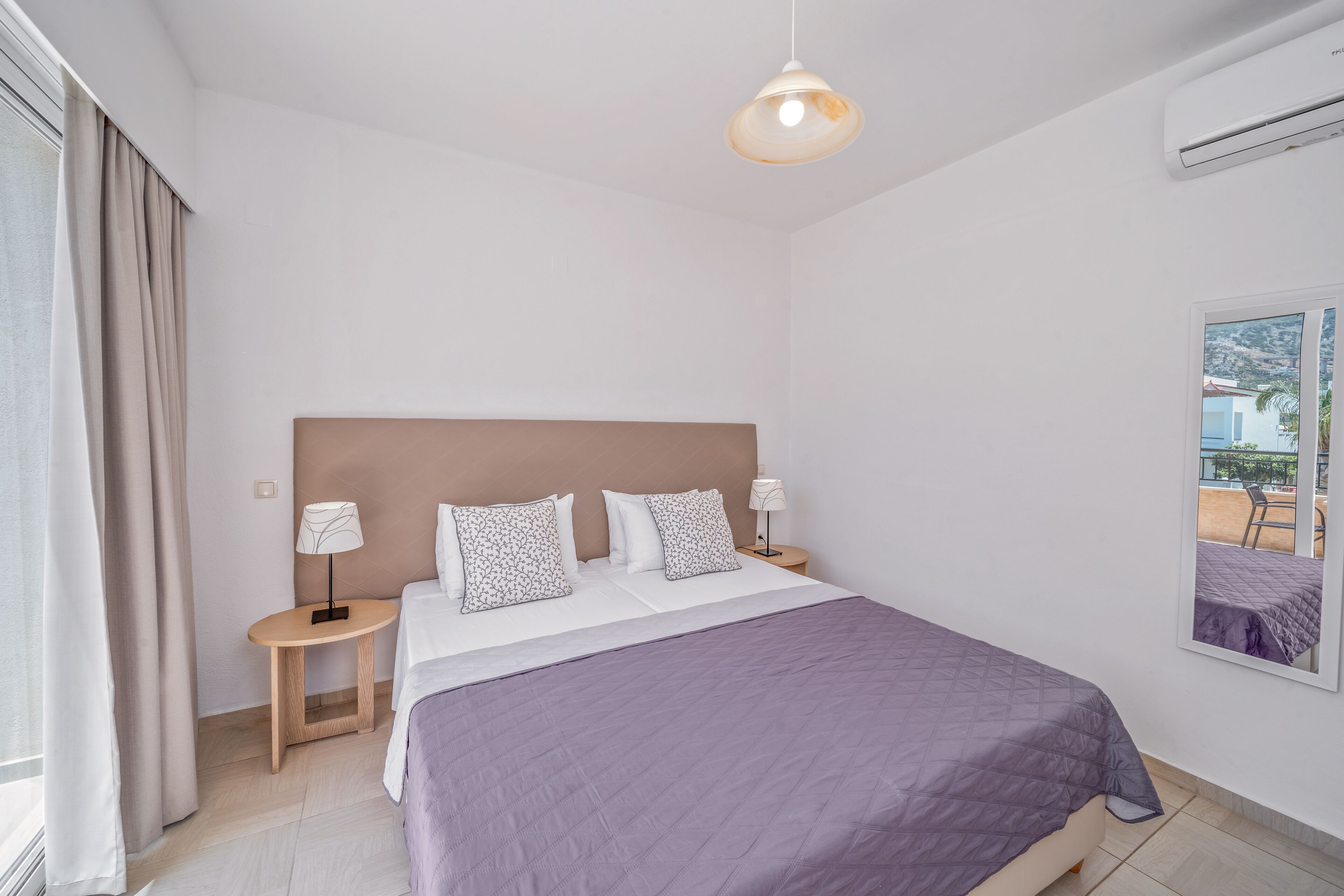 A modern hotel room with a large bed, two bedside tables with lamps, a view of the outdoors through the window, and an air conditioning unit on the wall.