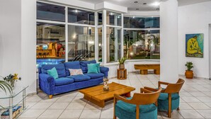 A spacious living area with a blue sofa, wooden coffee table, and teal chairs. Large windows offer views of the kitchen and dining area. - Maria Rousse (Hersonissos)