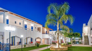 A multi-story hotel with a courtyard featuring palm trees and a well-maintained lawn. - Maria Rousse (Hersonissos)