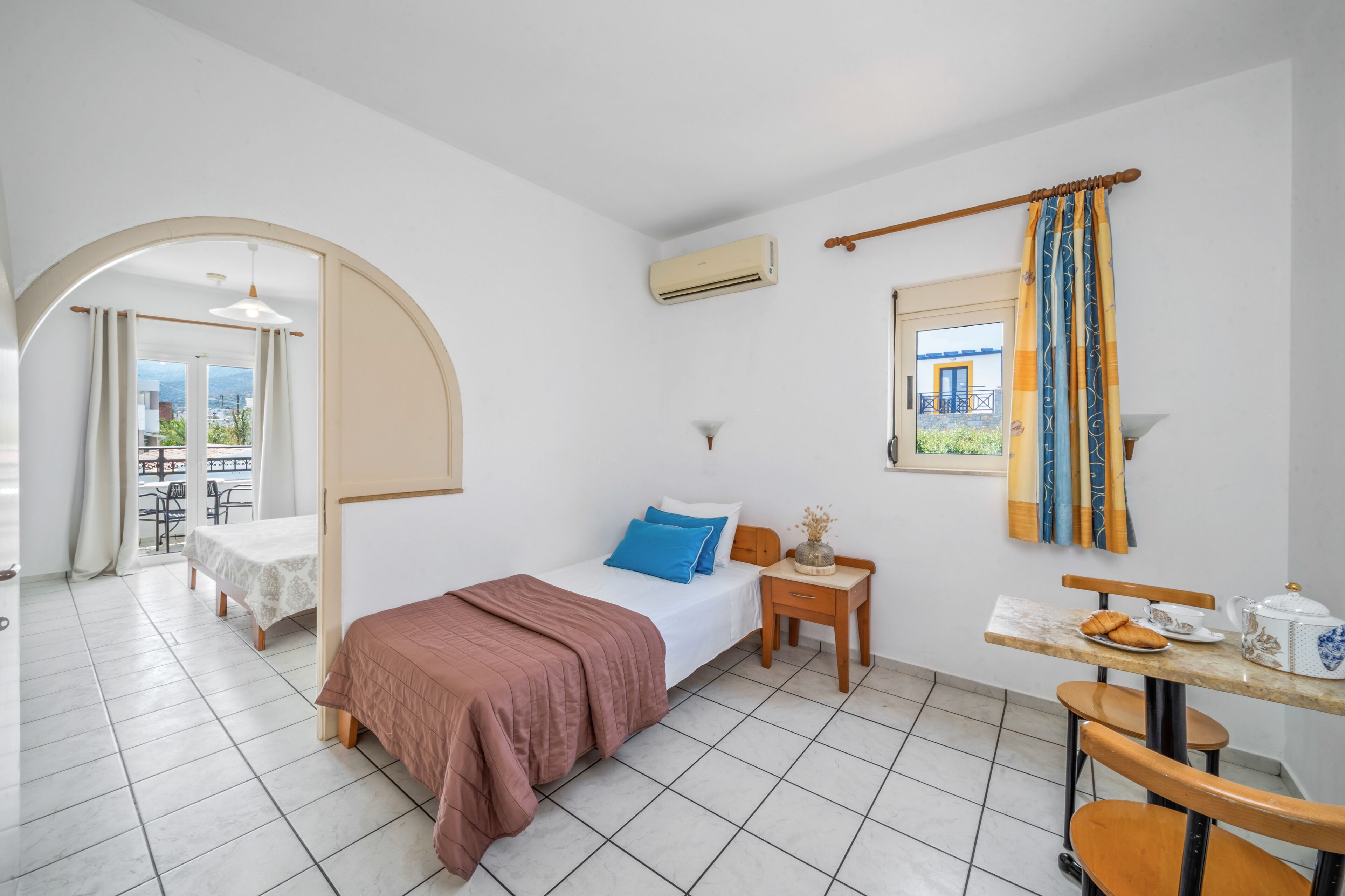 Une chambre d’hôtel avec un lit, une petite table sur laquelle se trouvent une tasse et des pâtisseries, une chaise, une fenêtre avec des rideaux et un appareil de climatisation.