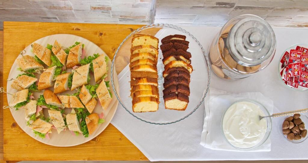 A table with a plate of sandwiches, a glass dish with layered cakes, a jar of cookies, and a bowl of cream with nuts.