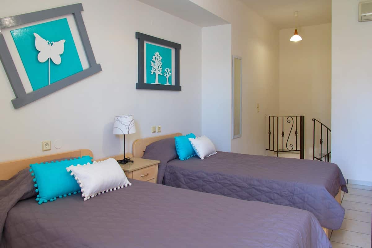 A bedroom with two beds, each with a purple bedspread and decorative pillows. There are wall art pieces above the beds, a bedside table with a lamp, and a door leading to another room.