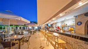 An outdoor bar area with tables and chairs, a counter with various bottles, and a stone wall. - Maria Rousse (Hersonissos)