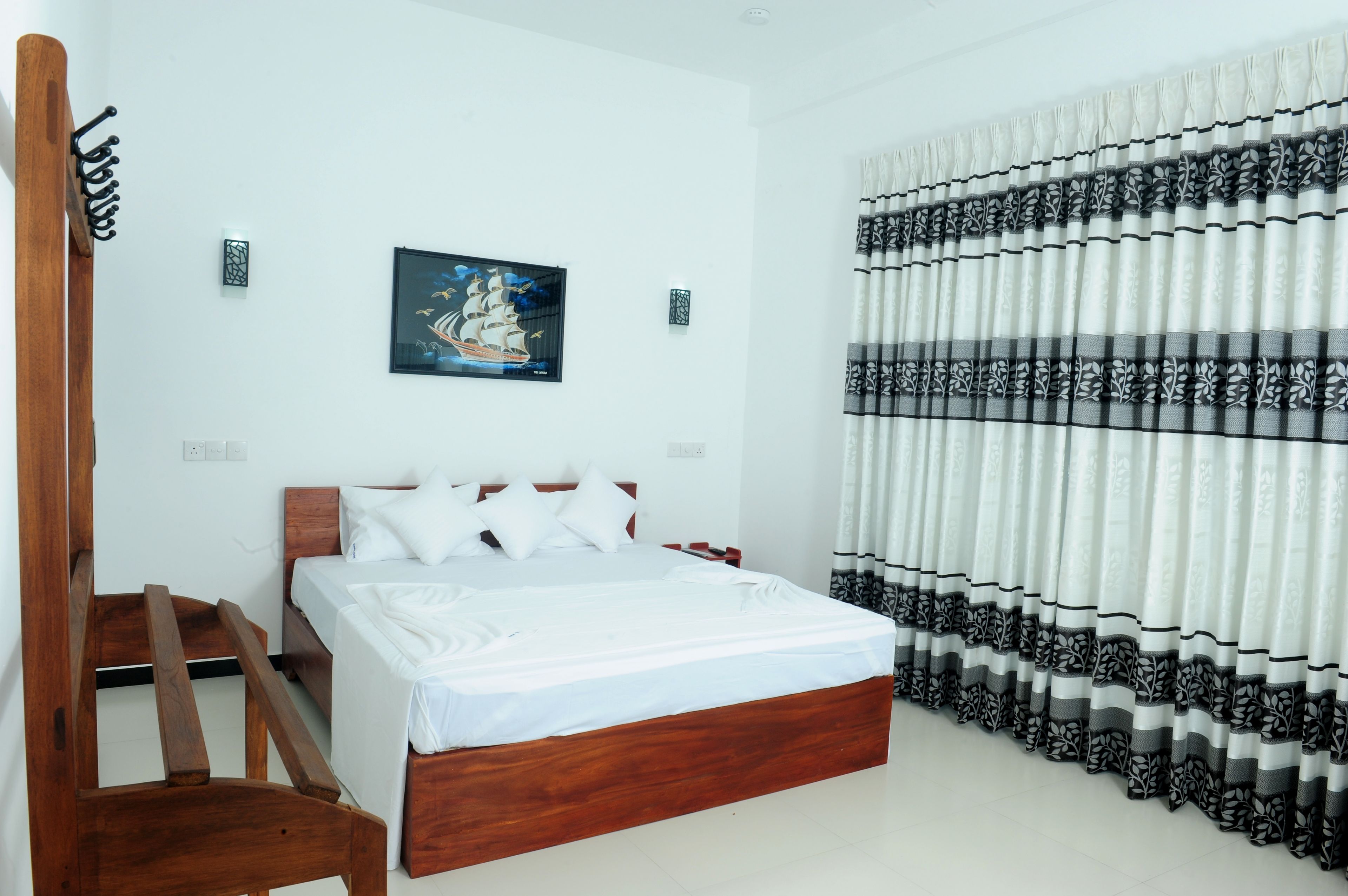A bedroom with a wooden bed, white bedding, a ship painting on the wall, and patterned curtains.